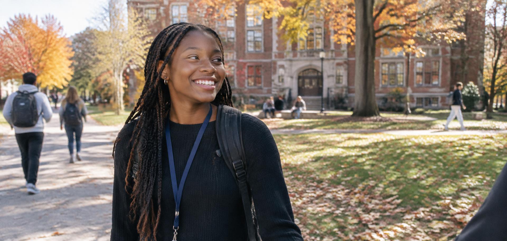 A smiling student with a backpack on a college campus quad in autumn